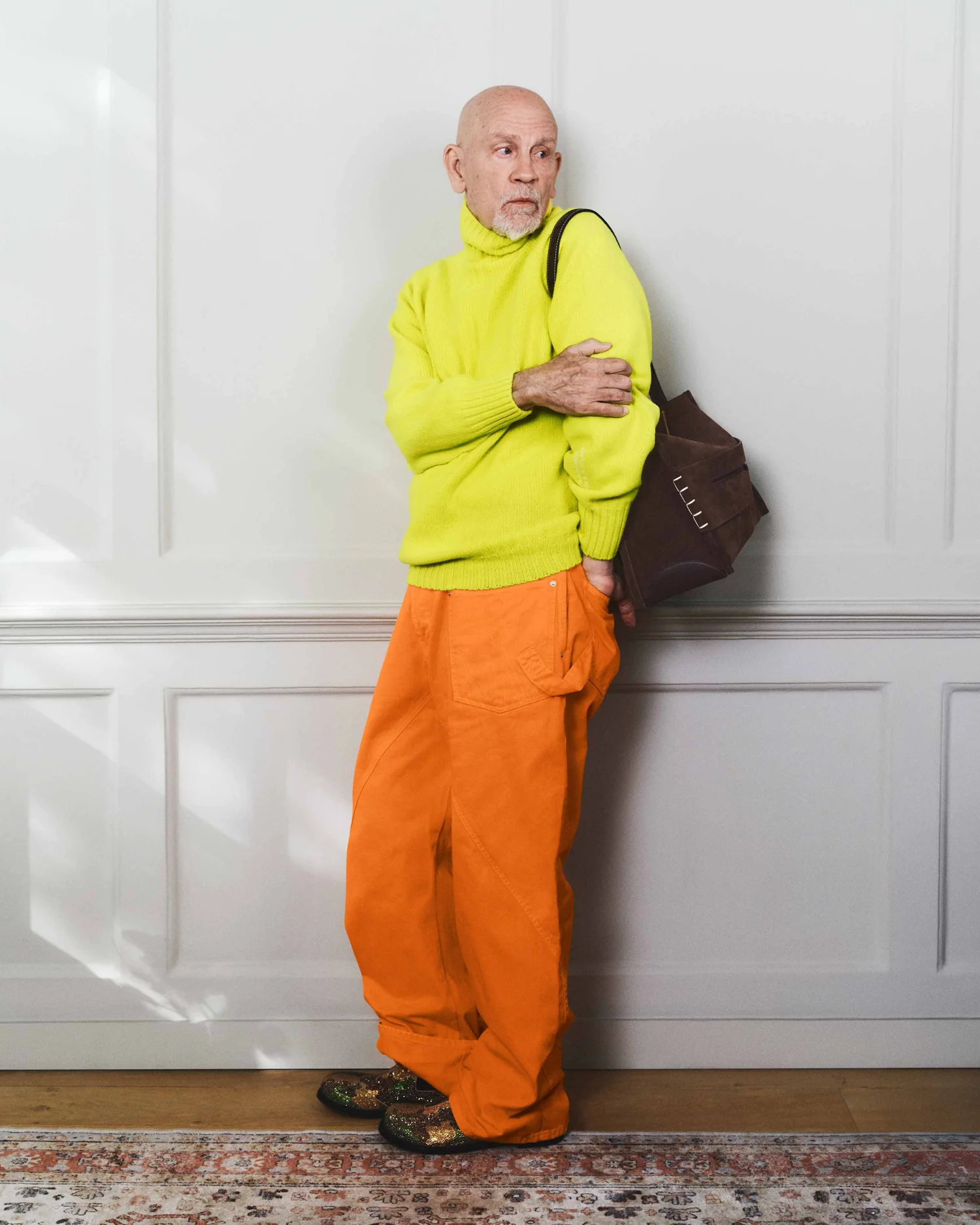 Glumac John Malkovic u naradžastim pantalonama, zelenom fluroscentnom džemperu sa braon torbom.