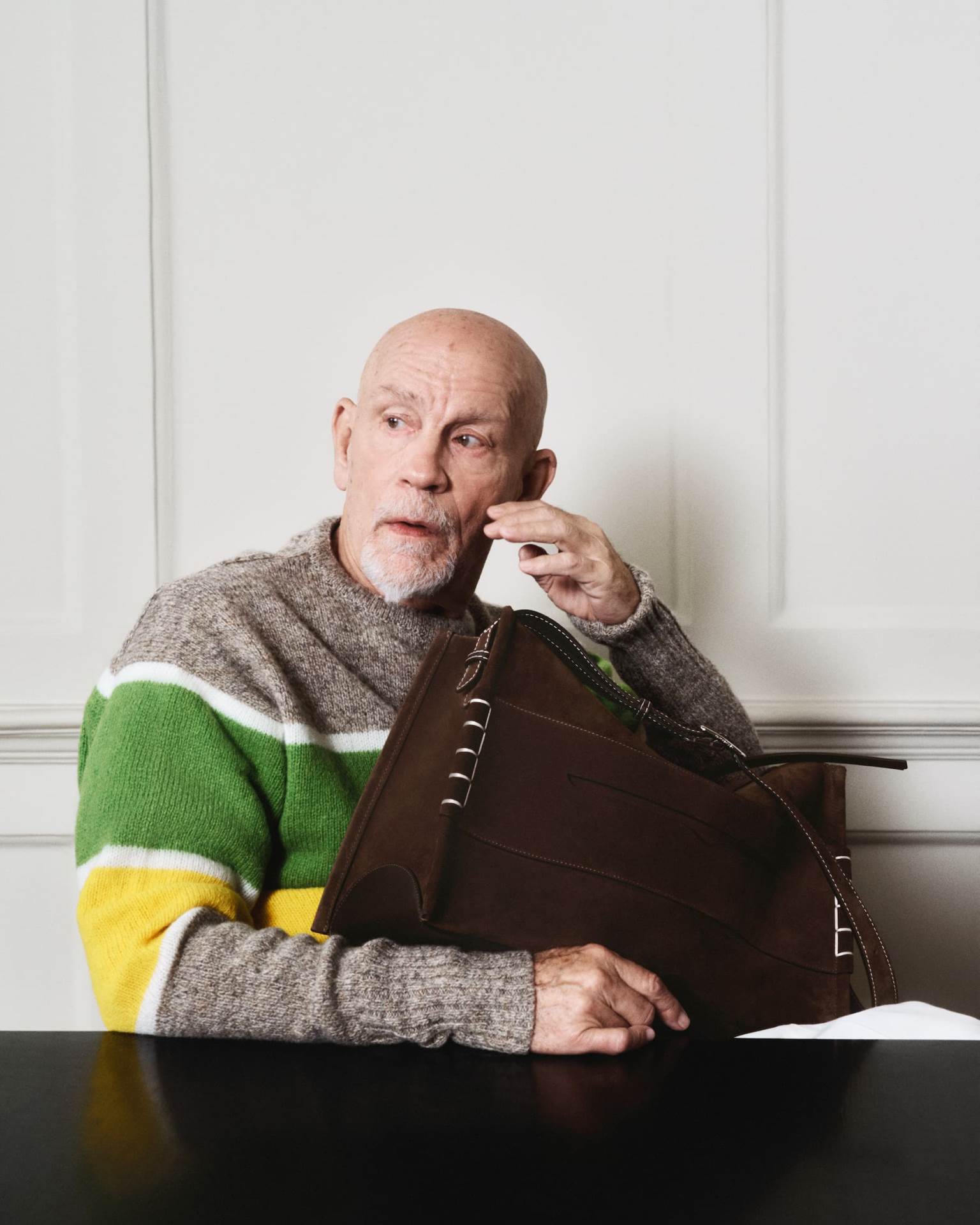 John Malkovich za kampanju brenda JW Anderson u sivom džemperu sa zelenom i žutom prugom i sa braon torbom.