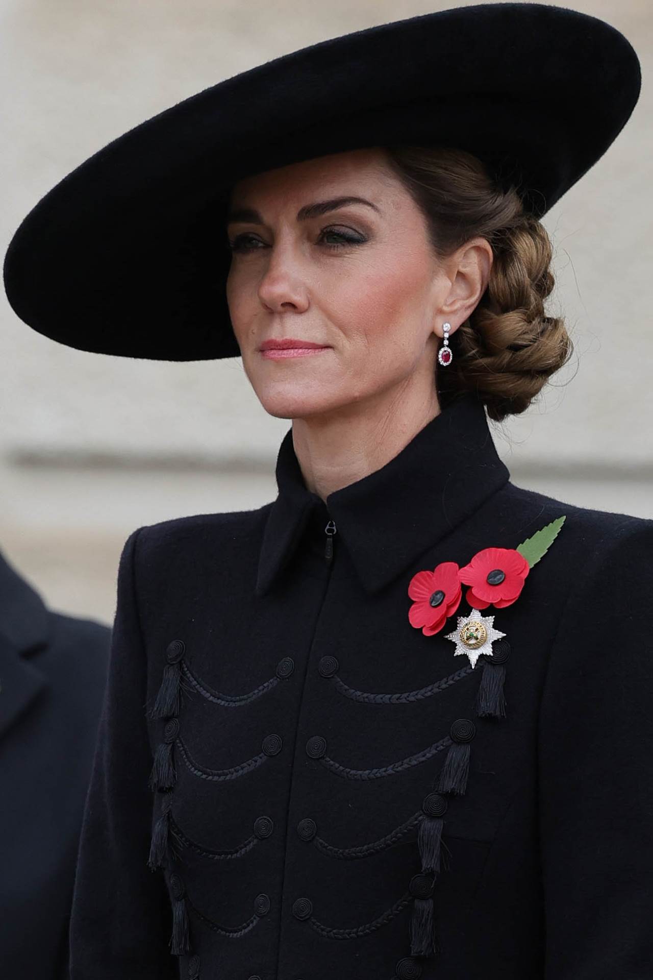 Kate Middleton u crnom coatdressu, sa šeširom iste nijanse i punđi sa strane.