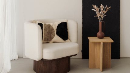 Stolica sa šarenim jastukom i stočić na kom je braon uska vaza sa cvetom.