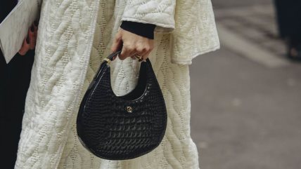 Žena u dugačkoj beloj jakni, crnim čizmama i sa crnom malom torbom.