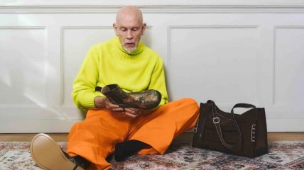 John Malkovich u narandžastim pantalonama, fluroscentnom džemperu, sedi na podu a pored njega je torba brenda JW Anderson.