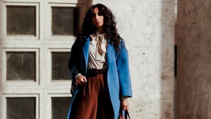 Žena u plavom kaputu, braon pantalonama i beloj košulji sa mašnom.