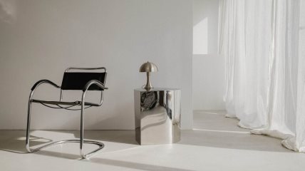 Srebrni pomoćni stočić sa metalnom lampom, pored crne stolice sa metalnim drškama.