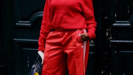 Žena u crvenom džemperu sa adiddas sportskim pantalonama, koje imaju tri linije sa strane, u istoj nijansi.