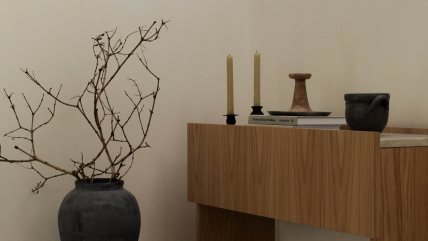 Drvena polica na kojoj su dve sveće, knjige i keremički ukrasi. Pored stoji vaza sa osušenim granjem.