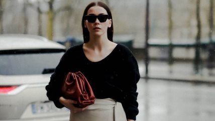 Žena u crnom džemperu i krem suknji, i sa crvenom torbom.