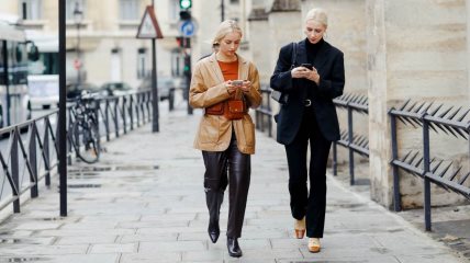Dve žene hodaju ulicom i gledaju u telefone. Jedna je u celokupnom black outfitu, druga u crnim pantalonama i braon jakni.