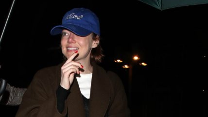Glumica Emma Stone u braon kaputu, beloj majici, sa plavim kačketom i crvenim manikirom.