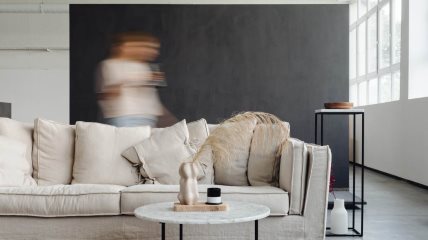 Žena u pokretu uslikana dok hoda iza krem kauča, ispred kog je stočić na kome je vaza.