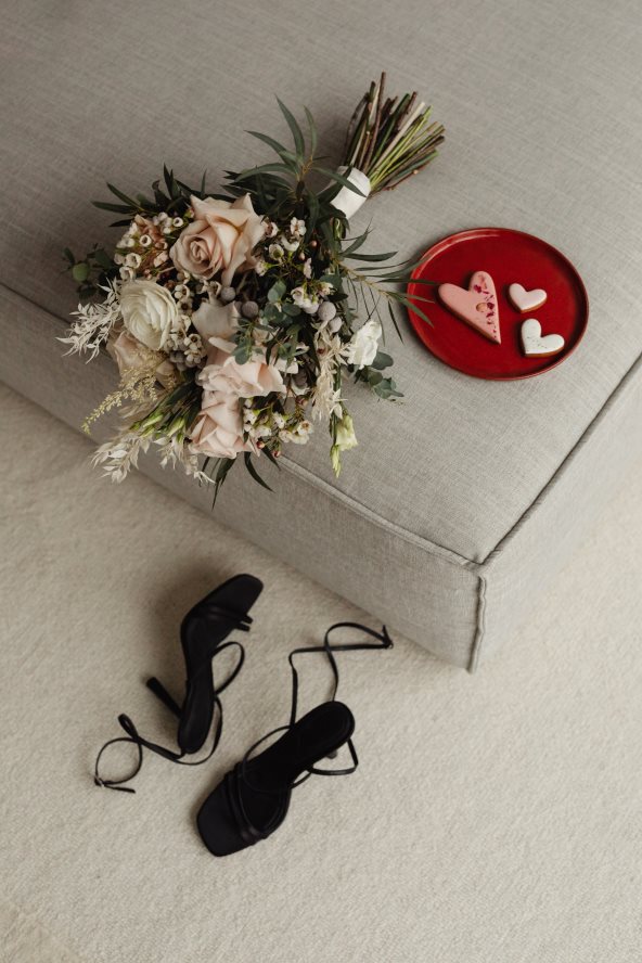 Na kauču stoji veliki buket cveća, crveni tanjir sa tri kolača u obliku srca, a na podu su crne sandale na štiklu.