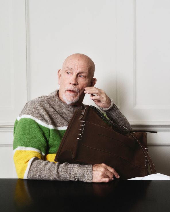 John Malkovich za kampanju brenda JW Anderson u sivom džemperu sa zelenom i žutom prugom i sa braon torbom.