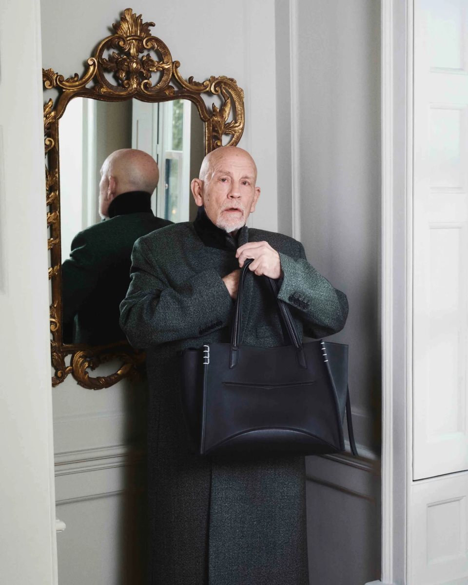 John Malkovich u sivom dugačkom kaputu sa crnom torbom.