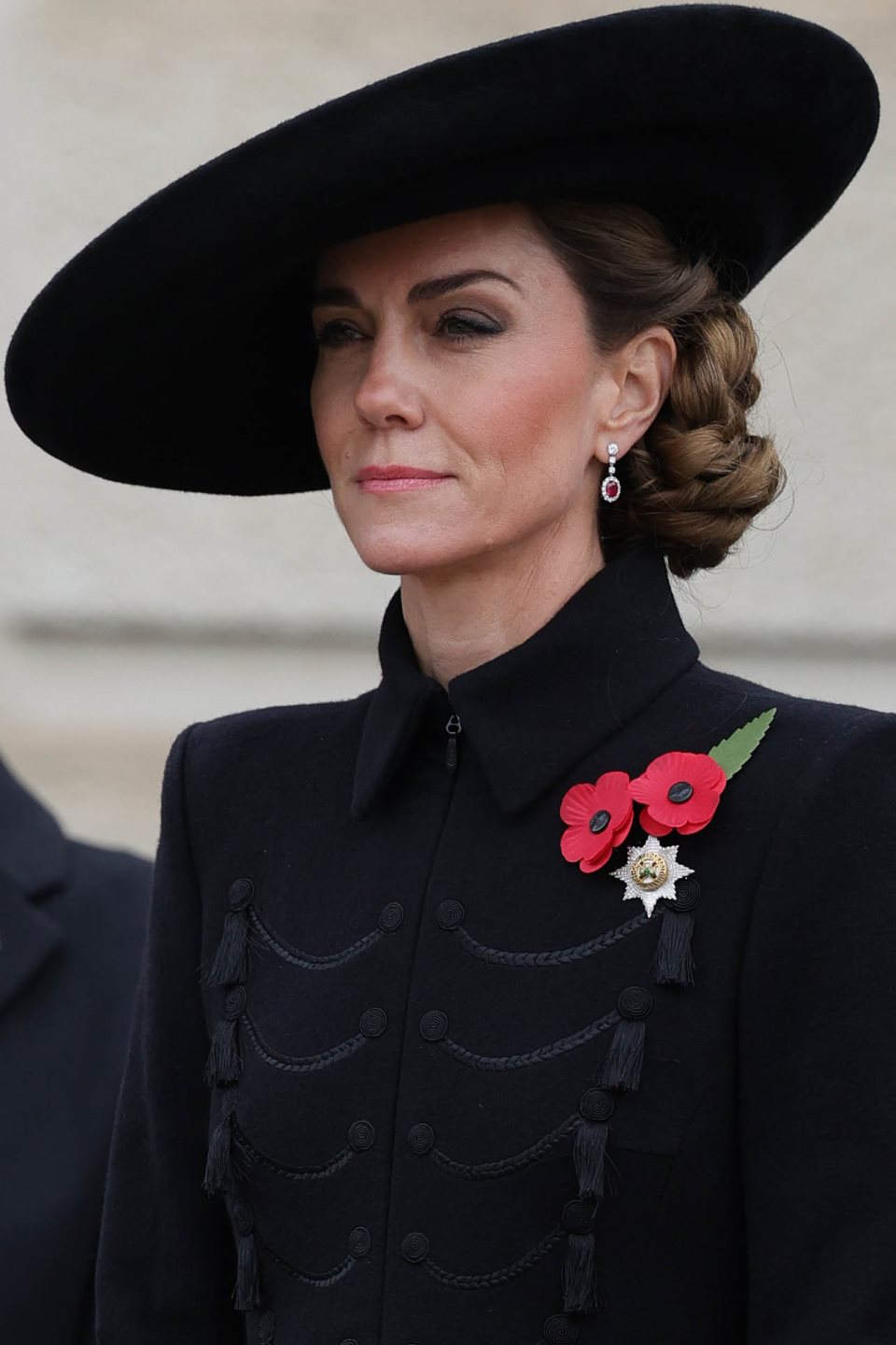 Kate Middleton u crnom coatdressu, sa šeširom iste nijanse i punđi sa strane.